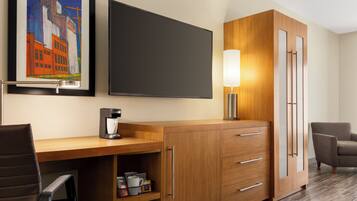 A hotel room with a wooden desk, a coffee maker, a chair, a cabinet with drawers, a framed picture on the wall, and a flat-screen TV.