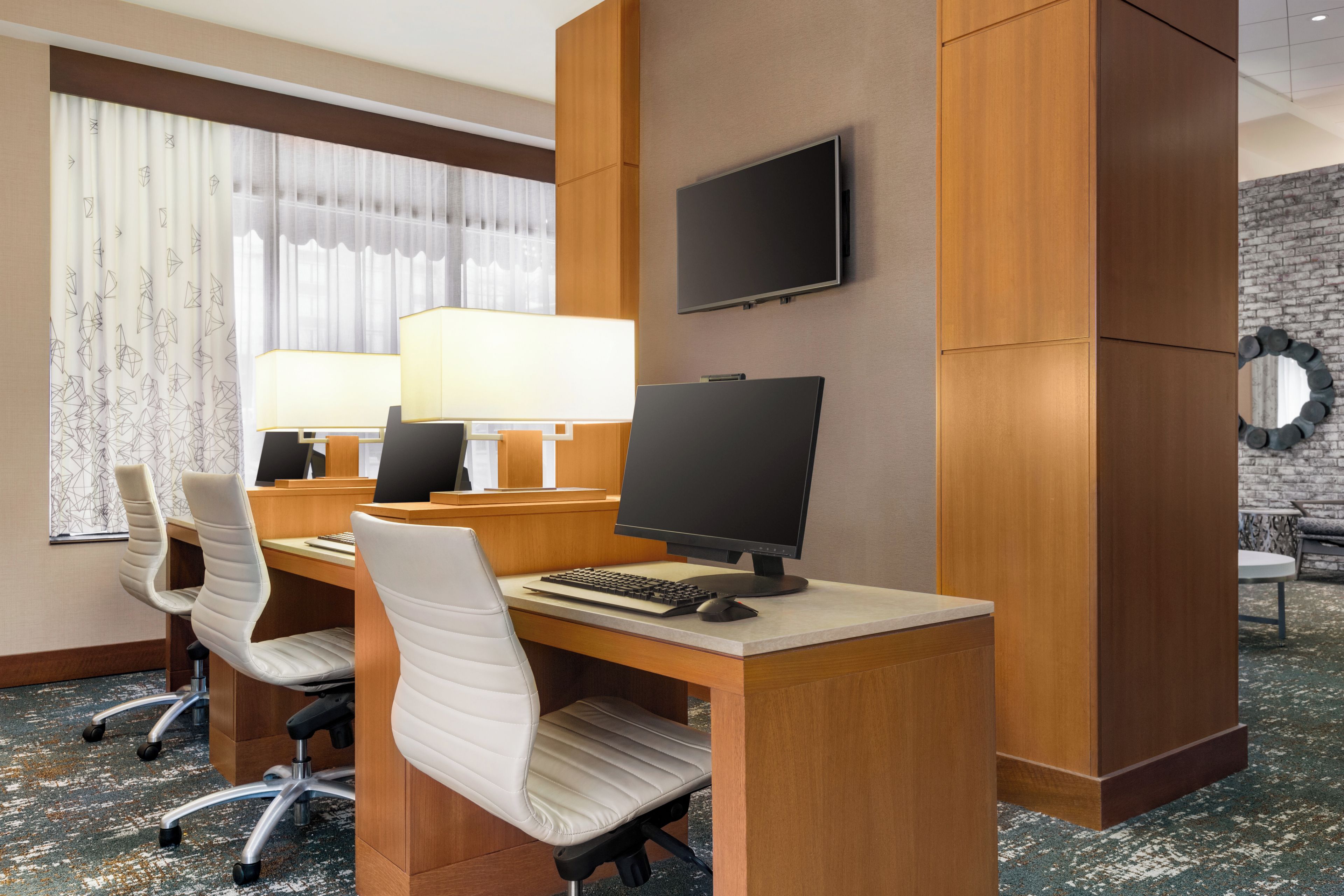 A hotel room with a desk, two chairs, a computer, a TV, and a window with curtains.