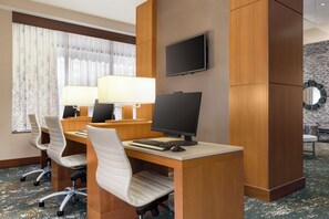 A hotel room with a desk, two chairs, a computer, a TV, and a window with curtains.