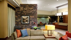 Eine moderne Hotellobby mit einer Ziegelfassade, einer Couch mit bunten Kissen, einem Tisch mit Lampe und einem Empfangsbereich im Hintergrund.
