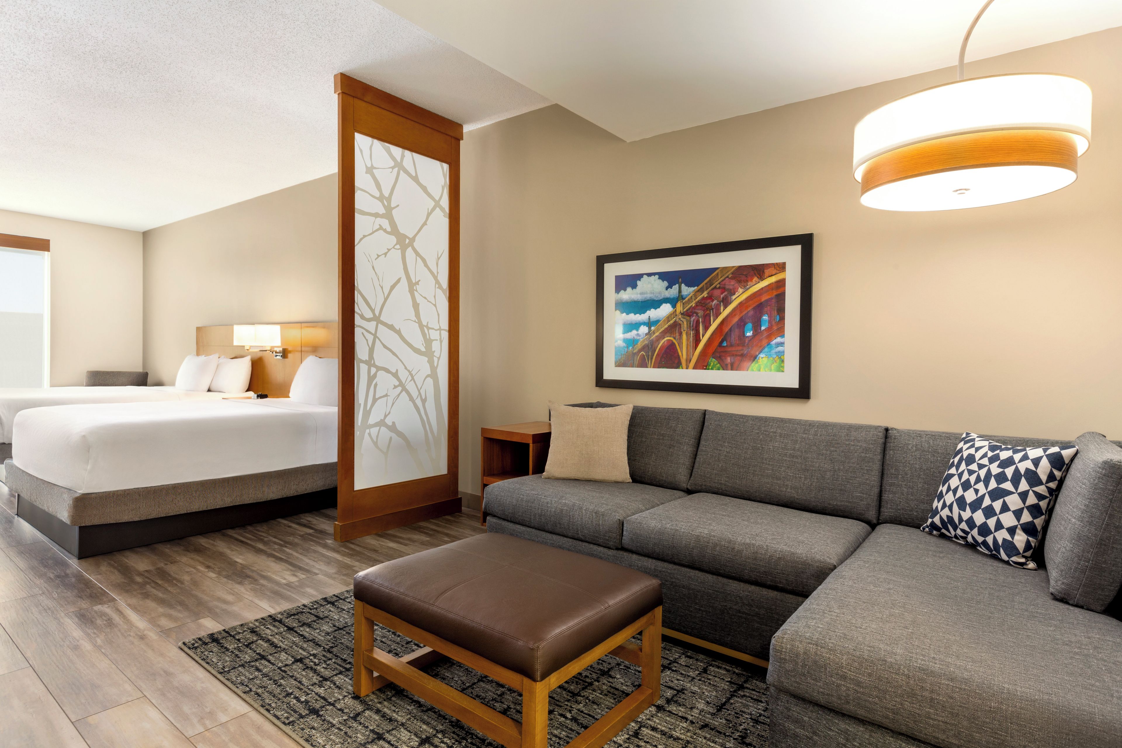 A hotel room with a bed, a sofa, an ottoman, a painting, and a ceiling light fixture.