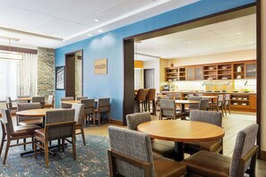 En hotelrestaurant med runde borde og stole, et køkken i baggrunden, og et skilt med teksten 'THE PLACERY'.