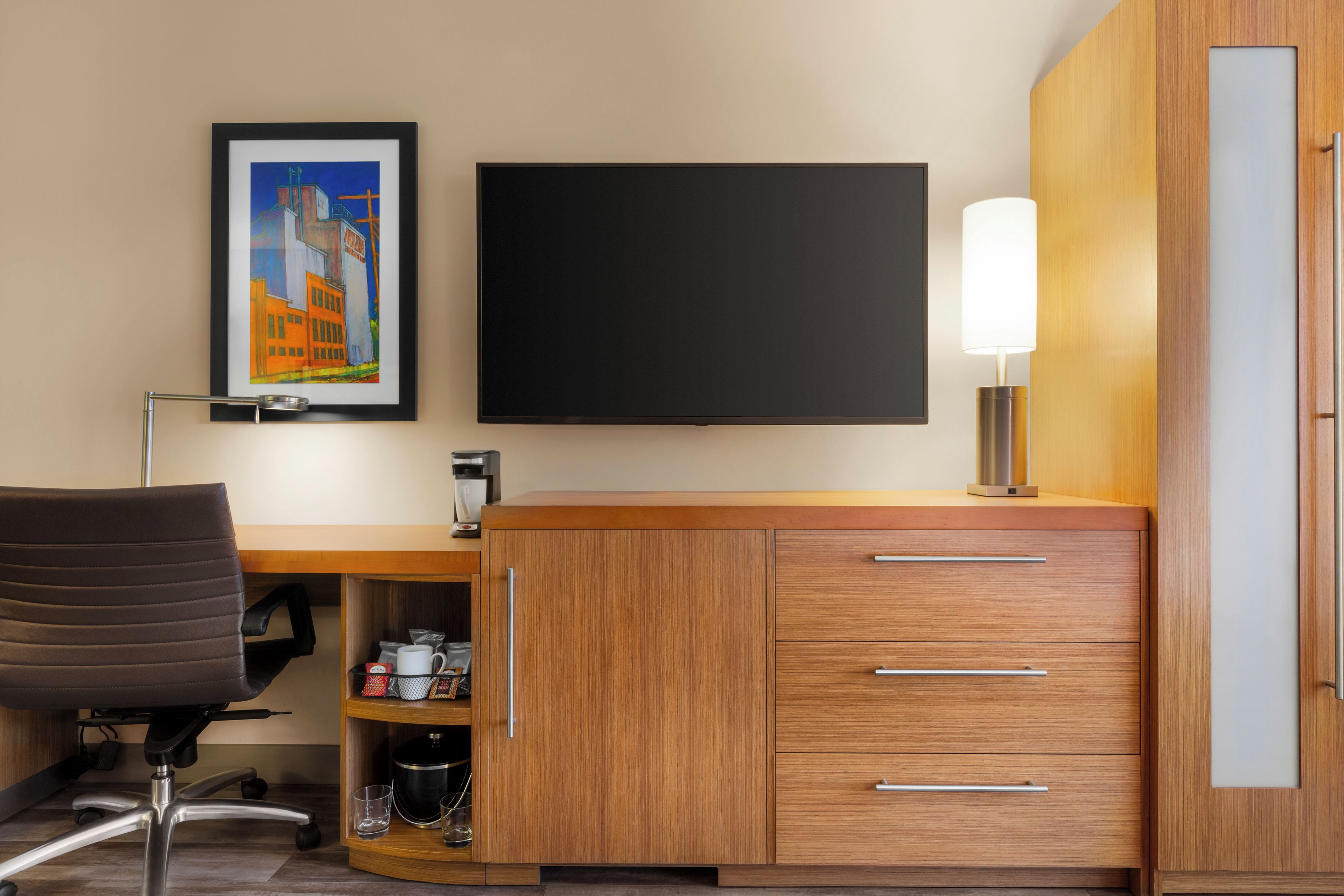 A hotel room with a wooden desk, a chair, a flat-screen TV, a lamp, and a framed picture on the wall.