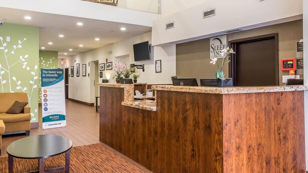 Un área de recepción de hotel con un mostrador de madera, un sofá y una mesa de centro. Se ven letreros de Sleep Inn y de Choice Rewards.