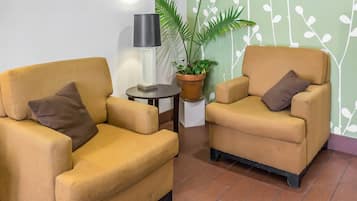 Two yellow armchairs with brown cushions, a small round table with a lamp, and a potted plant against a wall with a white leafy pattern.