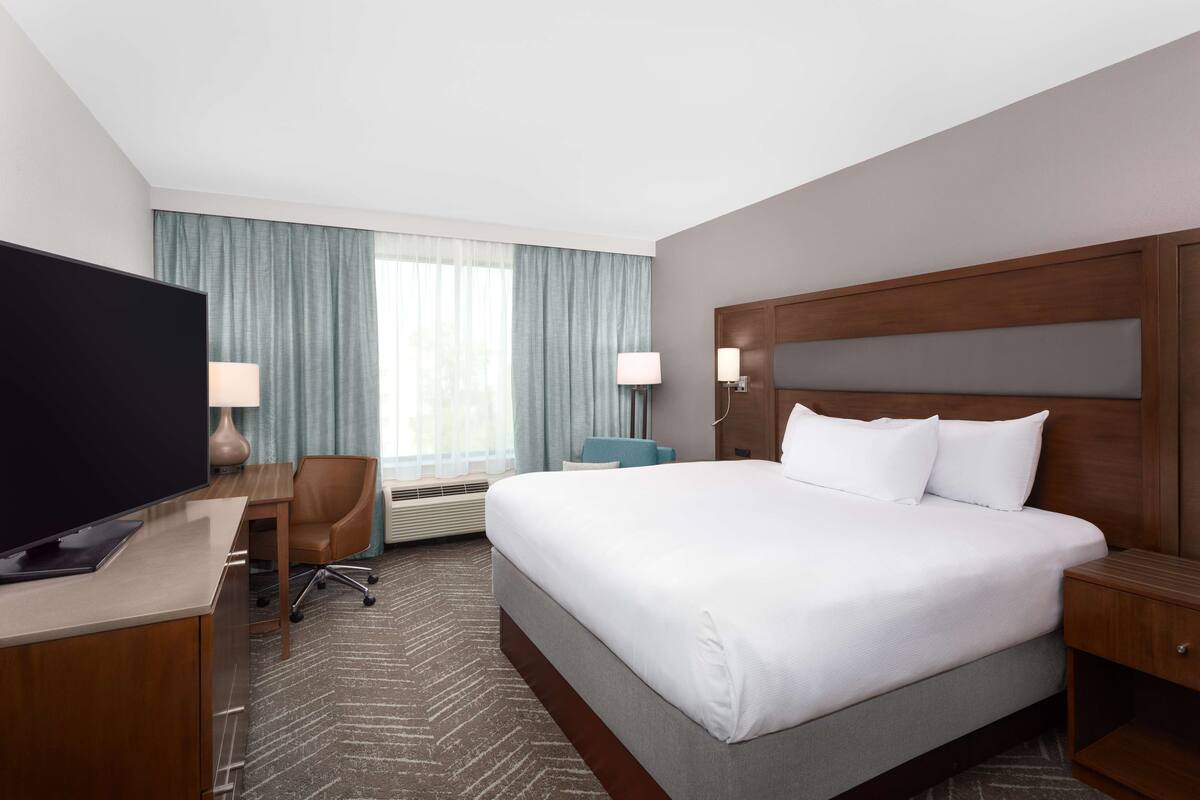 A hotel room with a large bed, a desk with a flat-screen TV, a chair, a desk lamp, and a window with curtains.