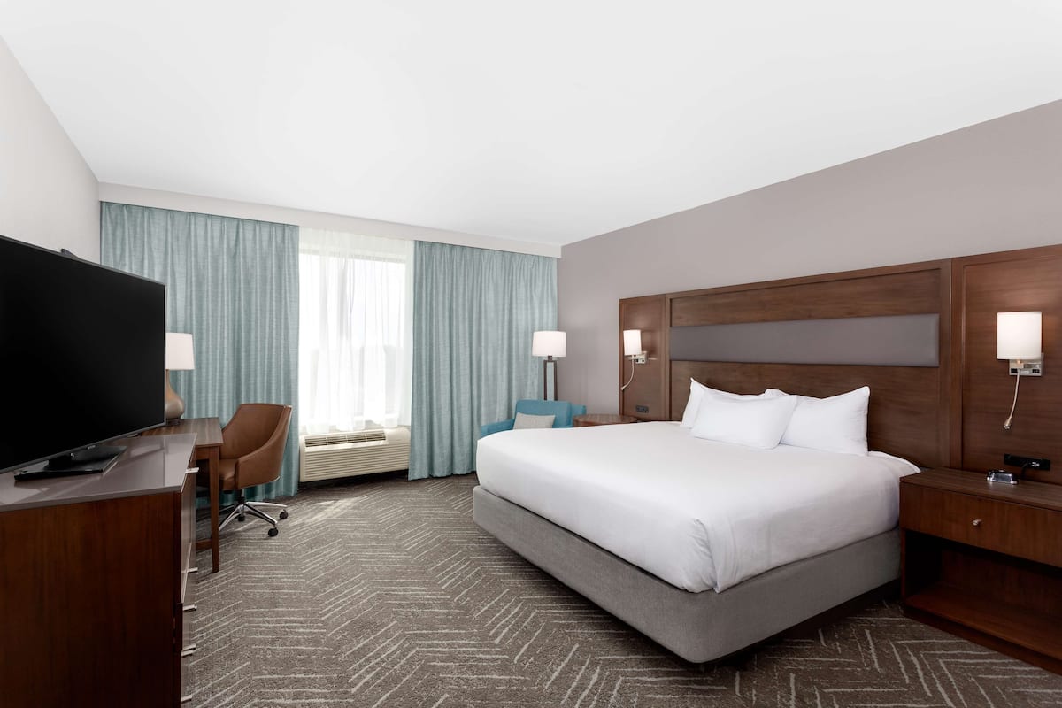 A hotel room with a large bed, a desk with a TV, a chair, and a window with curtains.