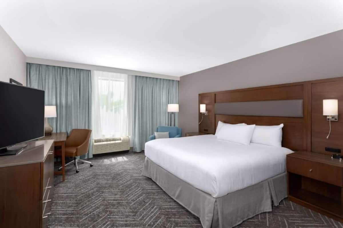 A hotel room with a large bed, a desk with a TV, a chair, a desk lamp, a nightstand, and a window with curtains.