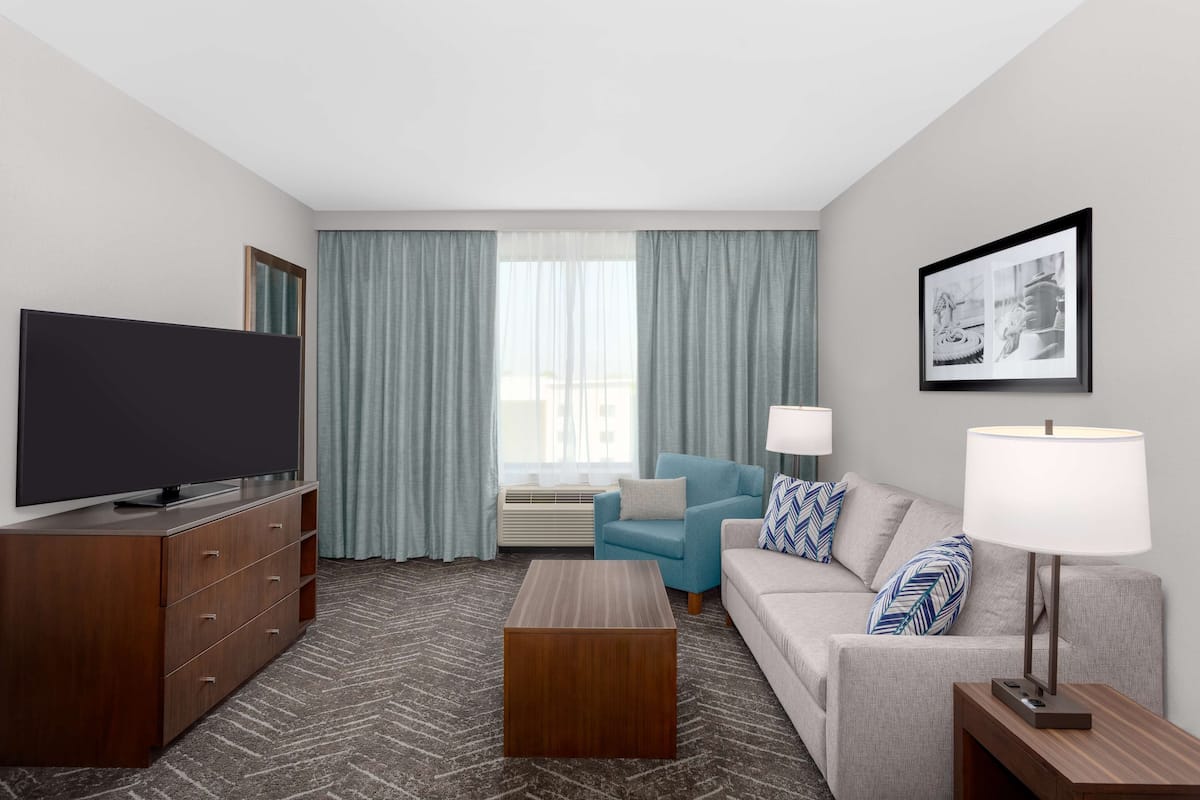 A hotel room with a flat-screen TV, a wooden cabinet, a coffee table, a sofa, a blue armchair, a lamp, and a framed picture on the wall.