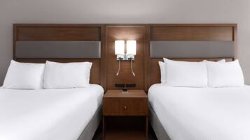 A hotel room with two beds, a wooden headboard, a nightstand with a lamp, and a small drawer.