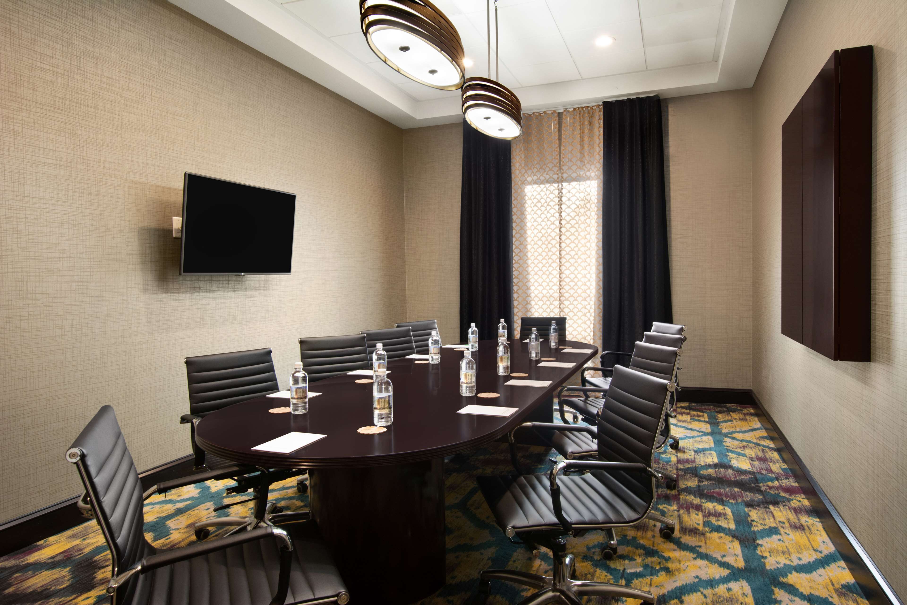 A conference room with a dark wooden table, black leather chairs, a flat-screen TV, and a patterned carpet.