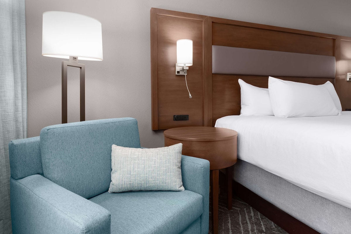 A hotel room with a bed, a blue armchair, a round wooden side table, and a wall-mounted lamp.