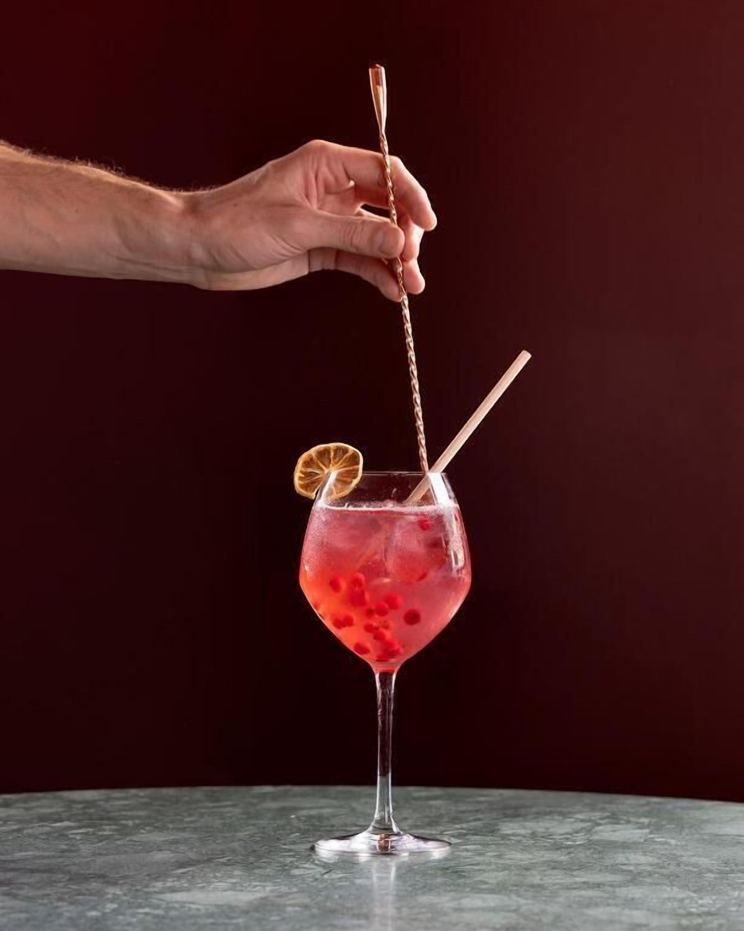 A hand placing a cocktail glass with a red drink, ice, and a slice of orange on a marble surface.