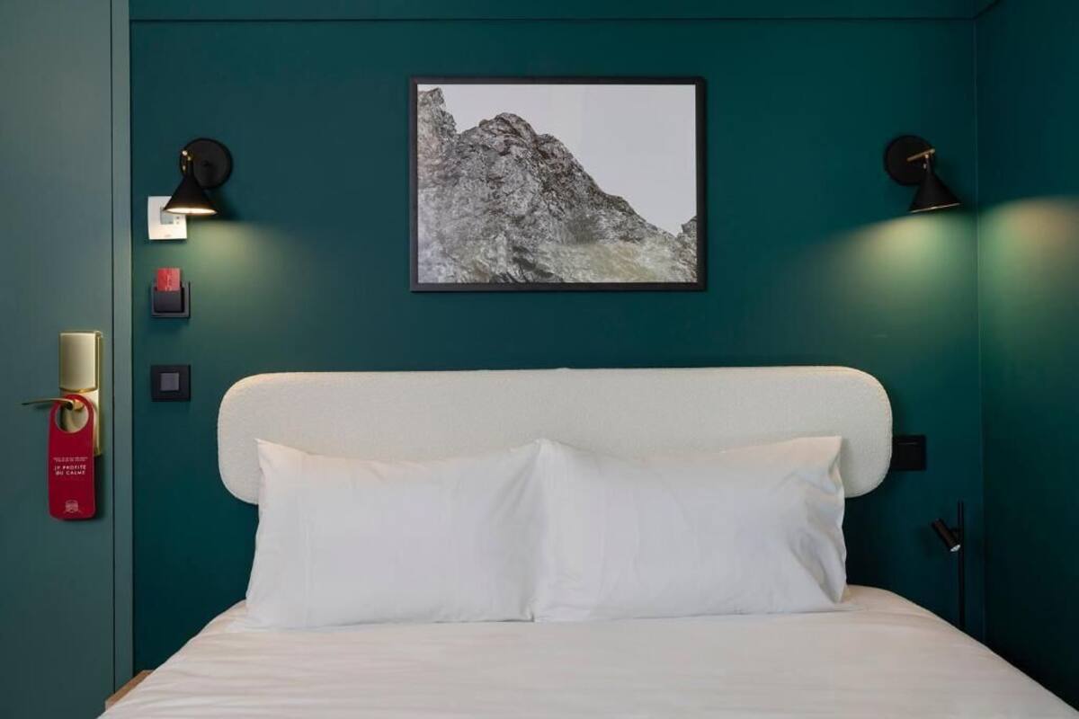 Une chambre d’hôtel avec un lit blanc, des murs de couleur turquoise et une photographie encadrée de montagnes au-dessus du lit.