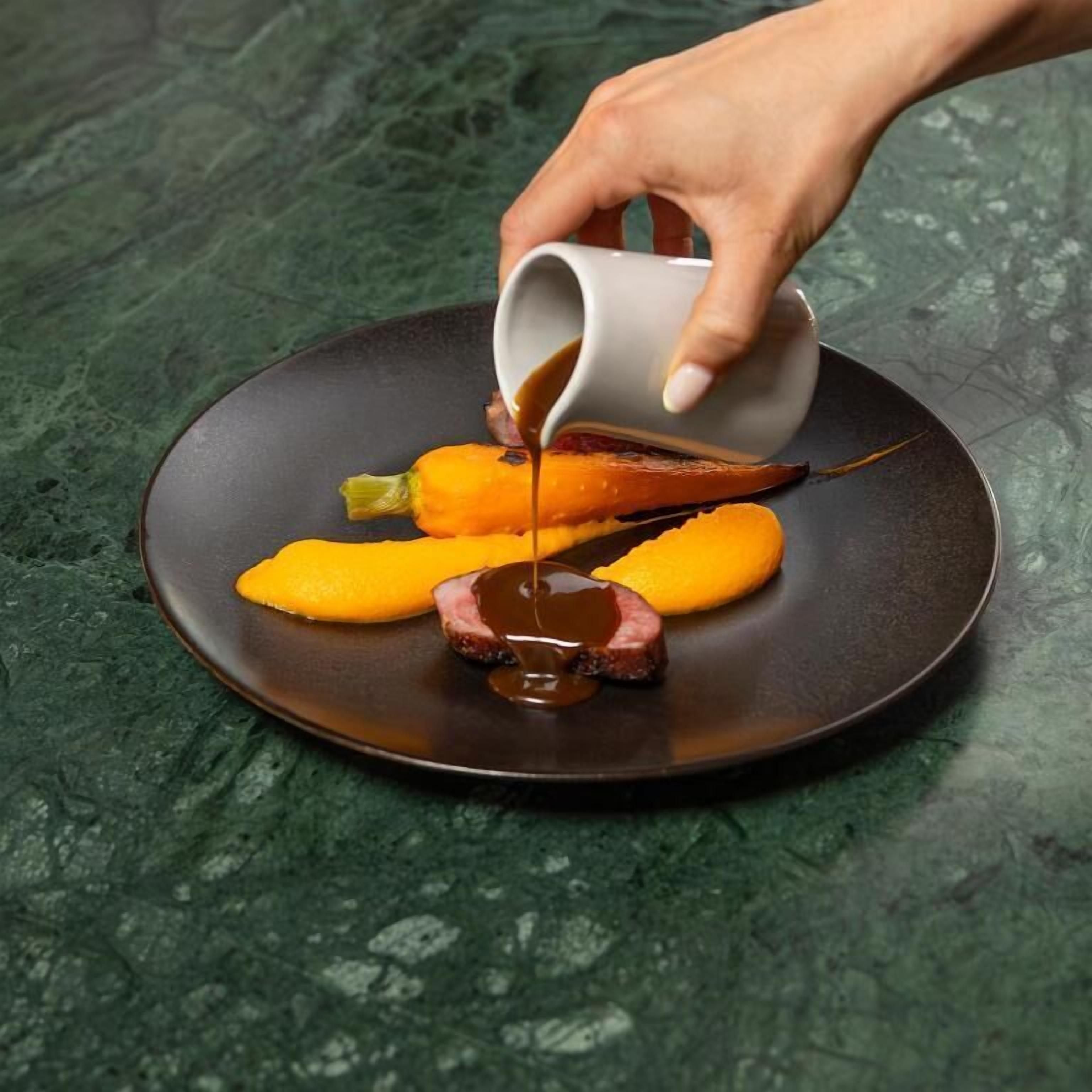 A hand pouring sauce over a dish with meat and vegetables on a plate.