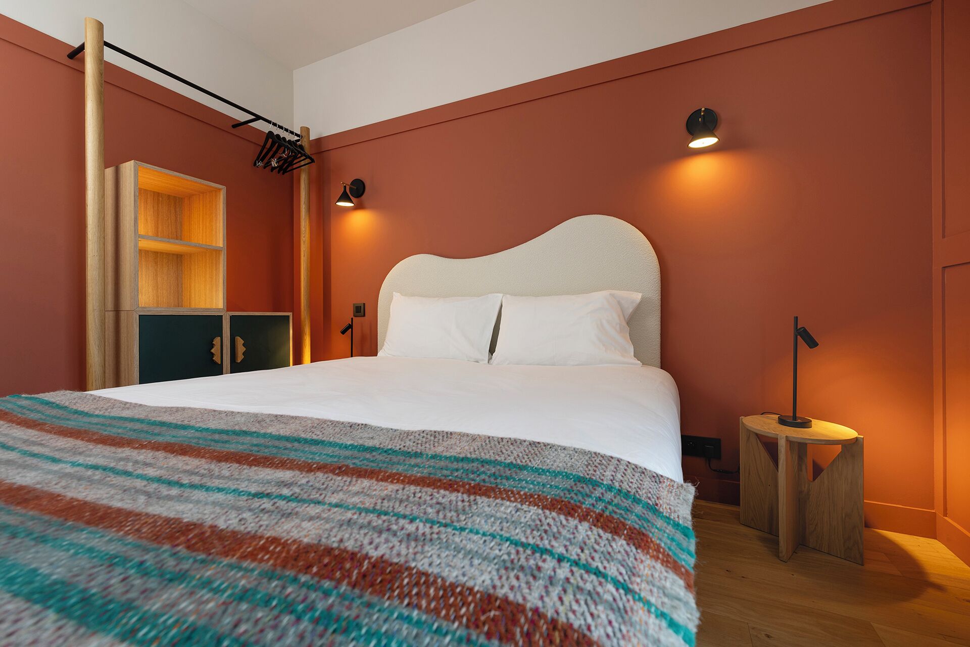 A bedroom with a large bed, a wooden wardrobe, a small table with a lamp, and a wall-mounted shelf.