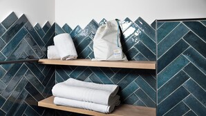 A bathroom with a herringbone tile backsplash, wooden shelves holding folded towels, and a white bag with a tag.