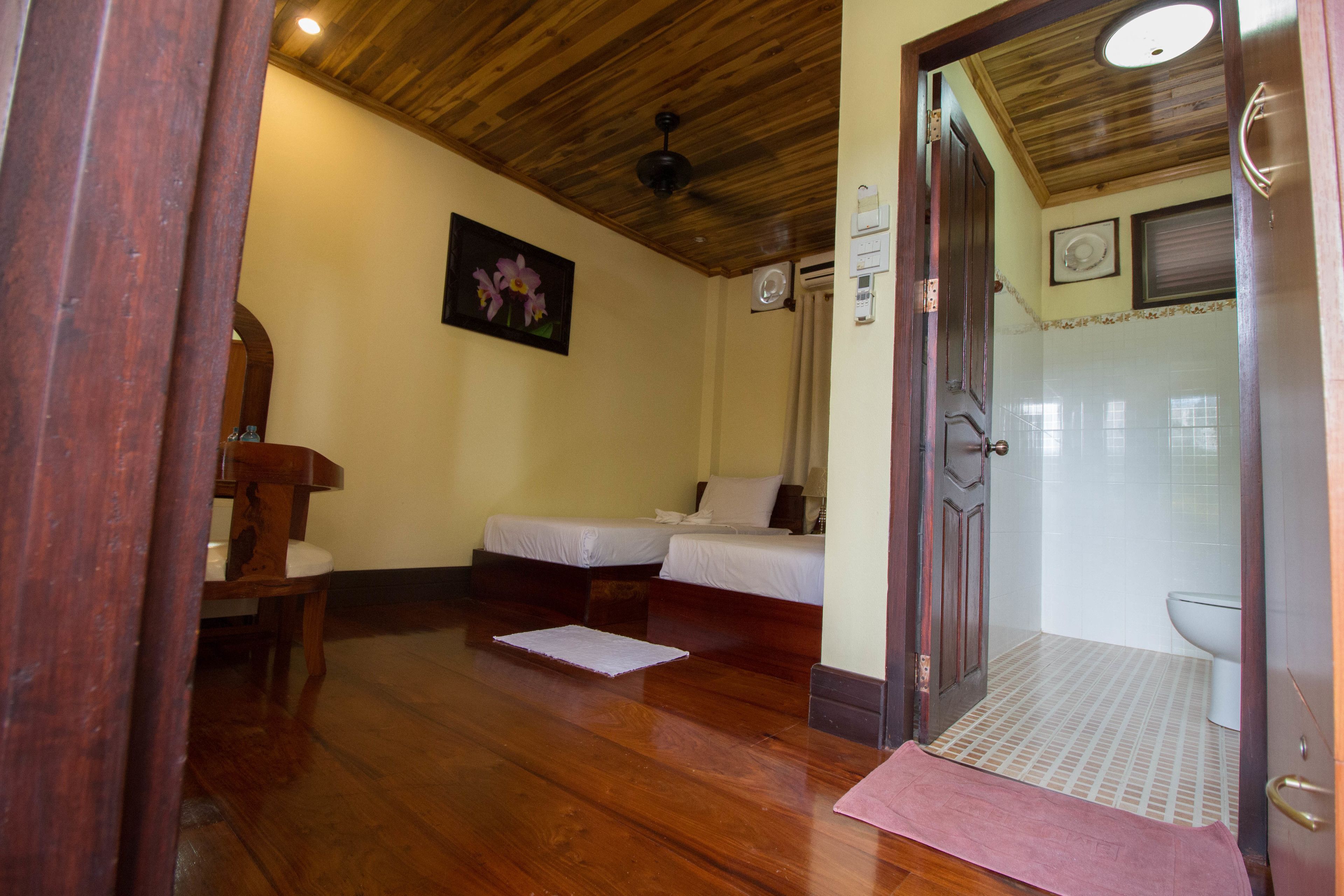 A room with wooden flooring, two beds with white linens, a wooden chair, a painting on the wall, a ceiling fan, and a bathroom with a toilet and shower.