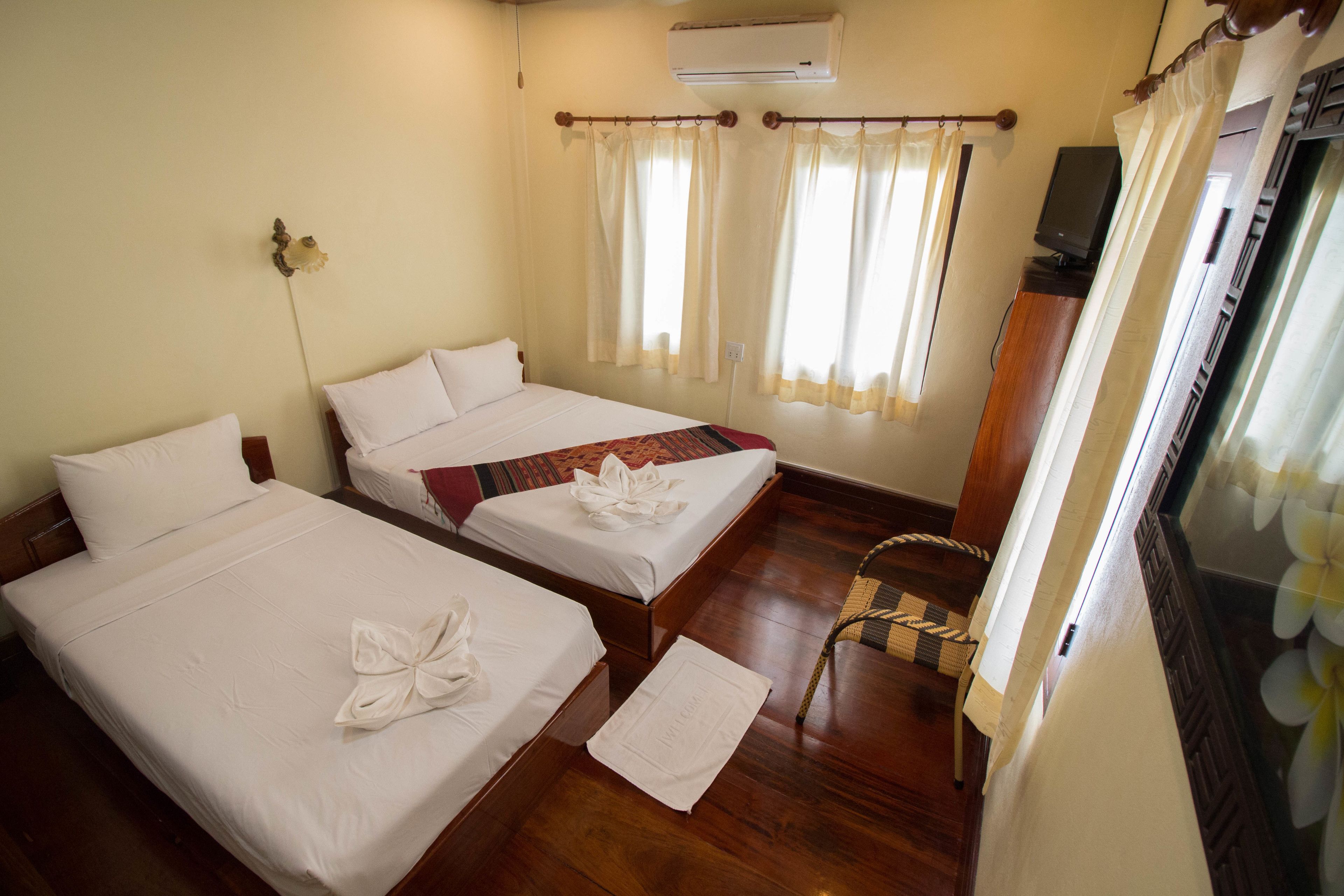 A hotel room with two beds, a wooden floor, an air conditioner, a television, and a window with curtains.