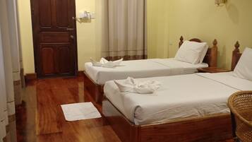 Quarto de hotel com duas camas de madeira, um ventilador de teto, uma porta de madeira e uma janela com cortinas.