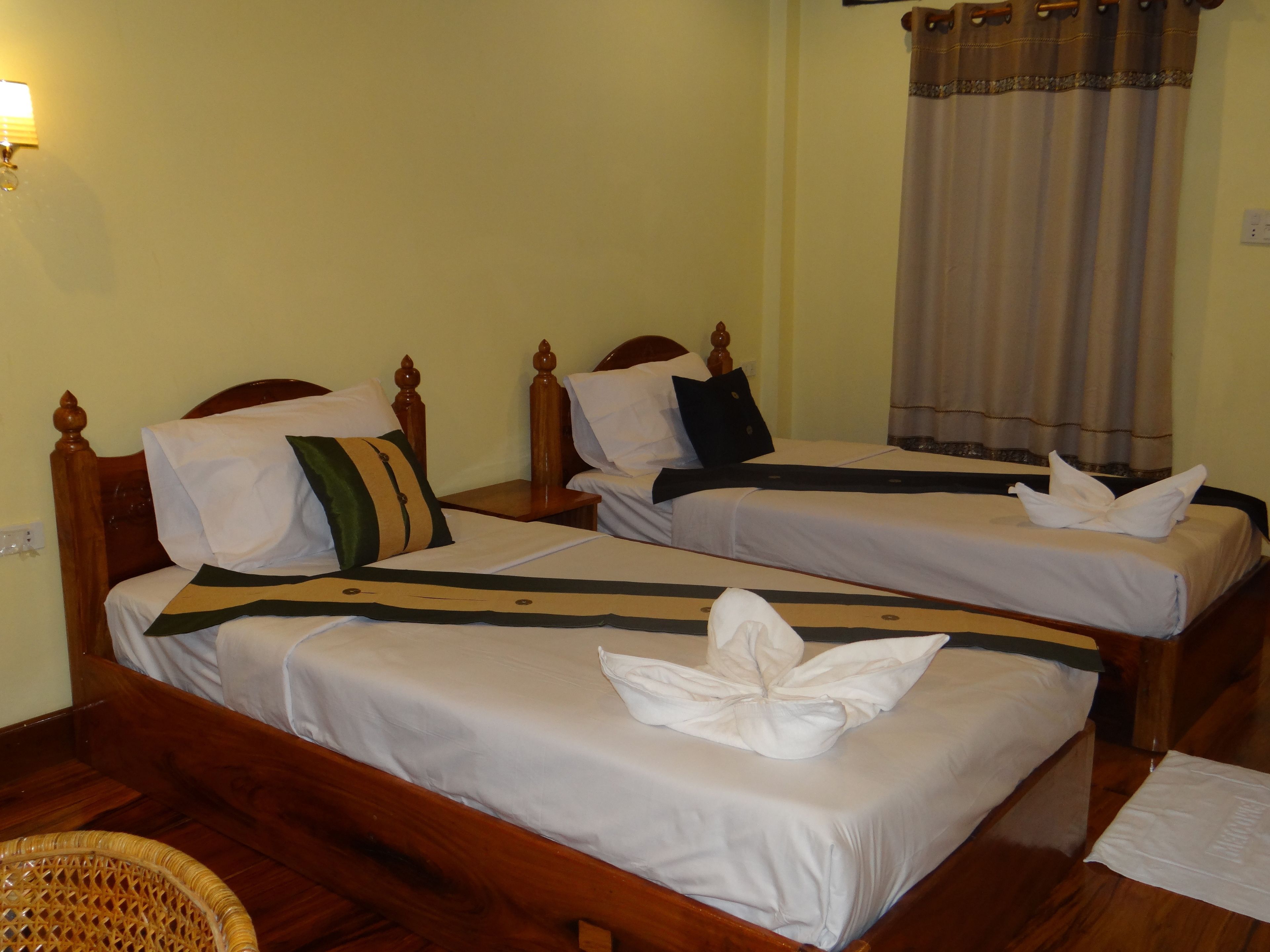 Habitación de hotel con dos camas de madera, cada una con cubrecama y almohadas blancas a juego. Hay una silla de mimbre en la esquina y una mesita de noche de madera con una lámpara.