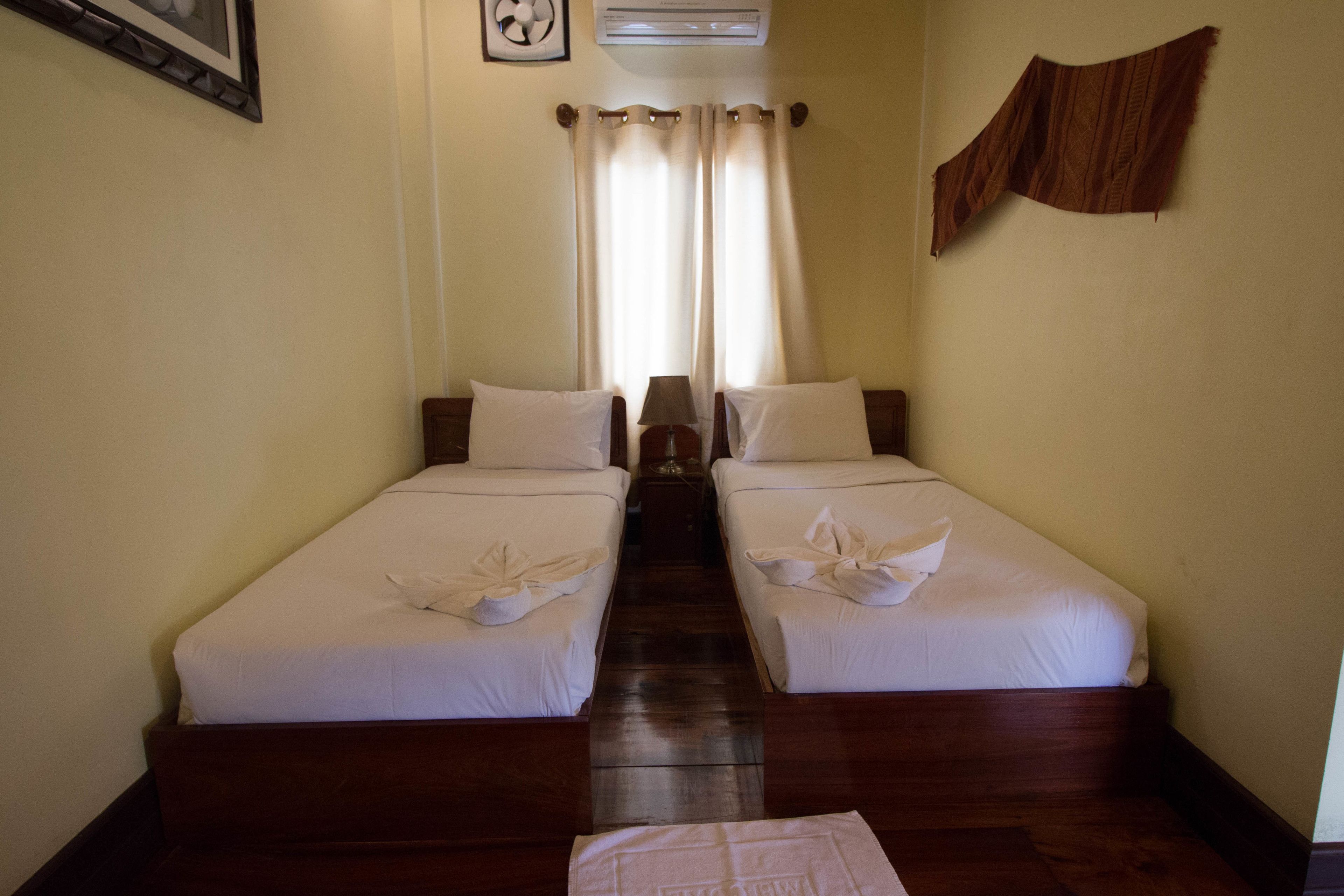 Une chambre d’hôtel avec deux lits simples, un plancher en bois, un ventilateur de plafond et une fenêtre avec des rideaux.