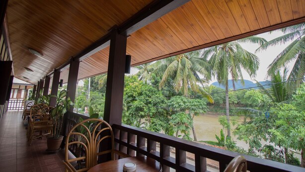 A covered outdoor seating area with wooden furniture and potted plants, overlooking a river and palm trees. - Villa Somphong (Luang Prabang)