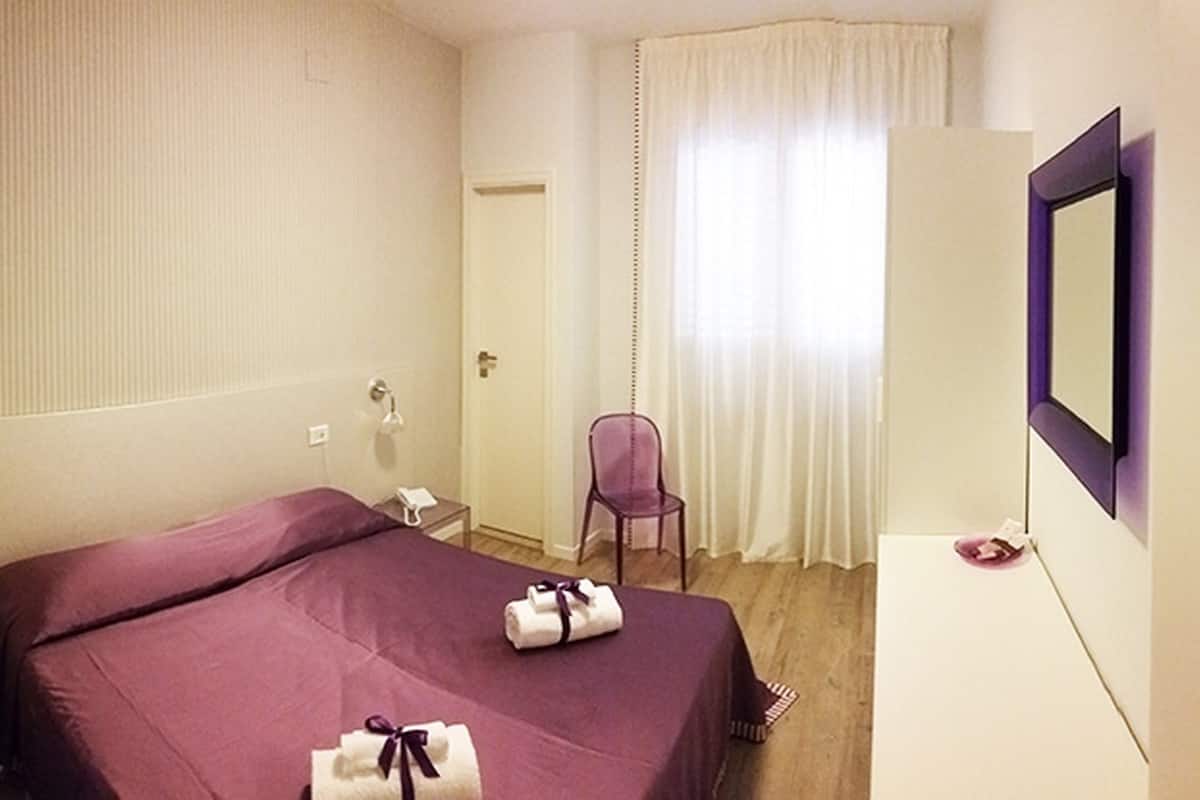 A hotel room with a bed, a chair, a desk, a mirror, and a door.