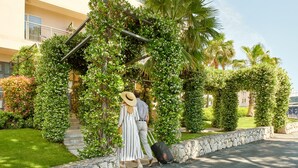 A couple walking through a lush, green garden area with a stone wall and steps leading to a building. - Dioklecijan Hotel & Residence (Split)