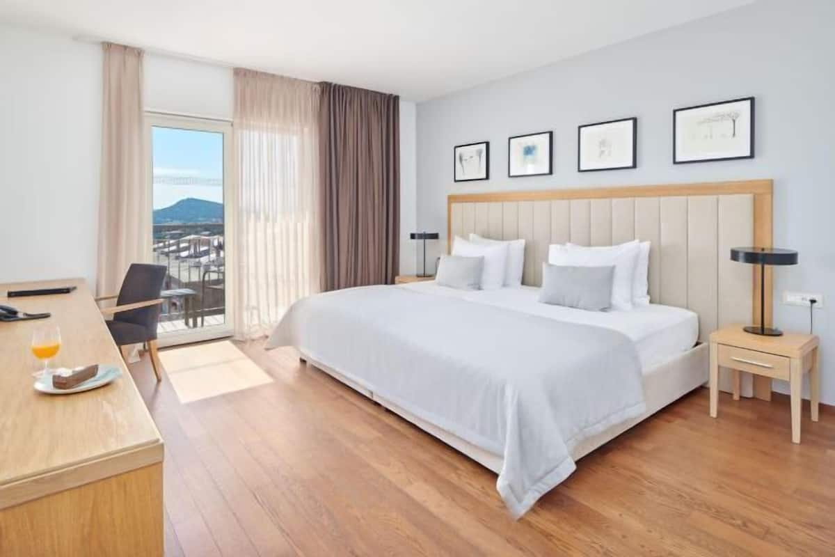Habitación de hotel con una cama grande, un escritorio con silla, una mesita de noche, una lámpara y vistas al exterior.