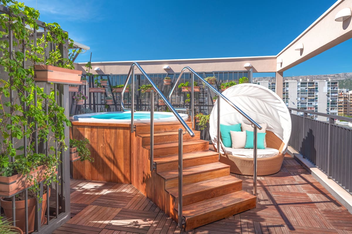 Terraza en la azotea con jacuzzi, escalones de madera y una silla redonda de mimbre con cojines.