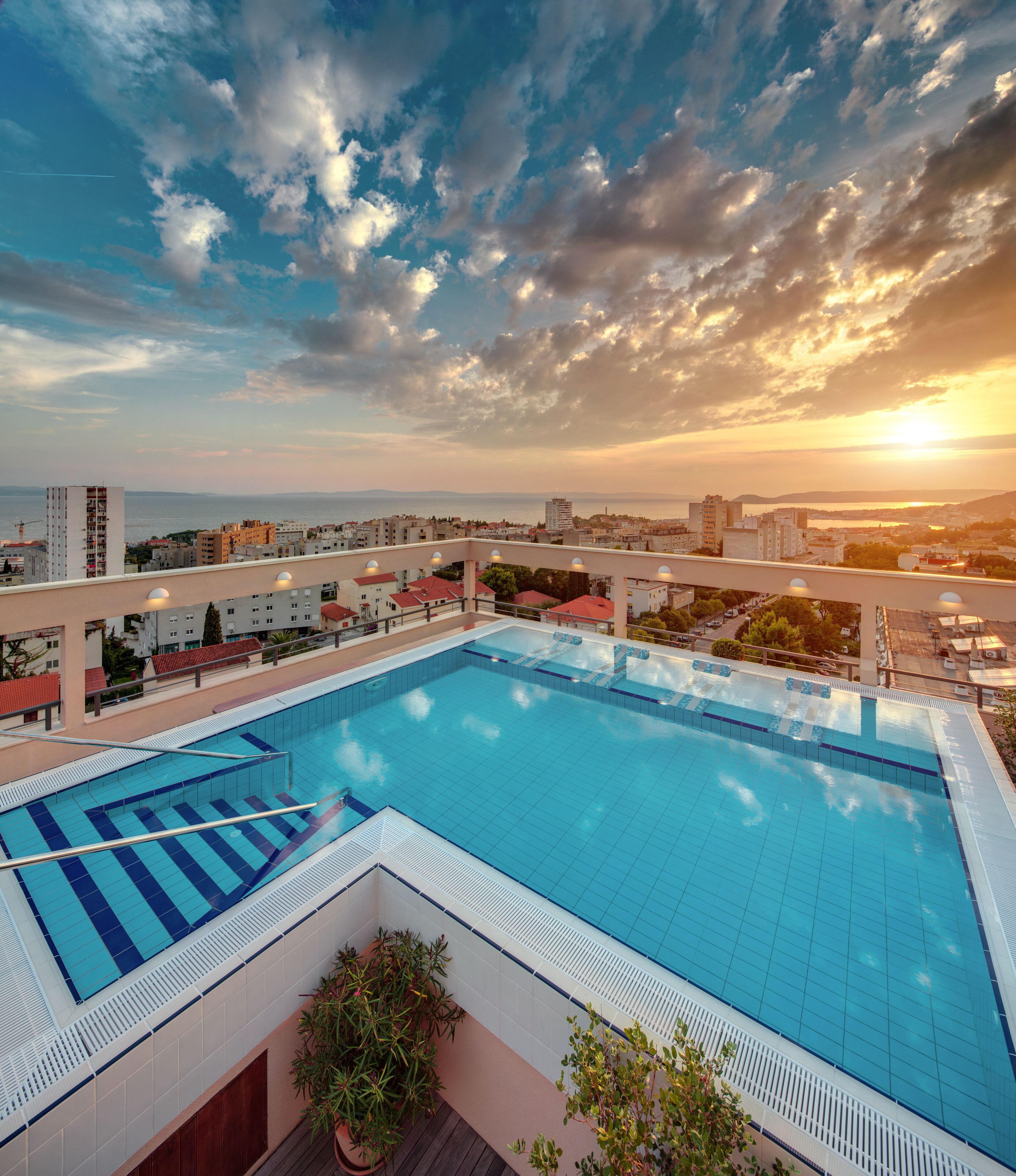 A rooftop swimming pool with clear blue water, surrounded by buildings and a sunset in the background.