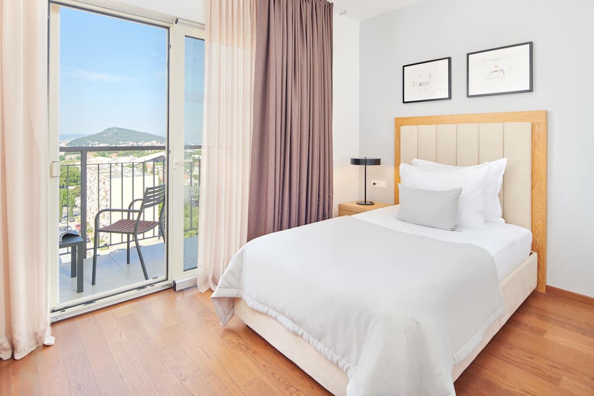 Habitación de hotel con una cama grande, una mesita de noche, una silla y vistas a un balcón con el paisaje urbano.