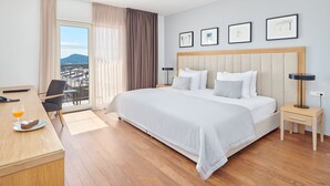 A hotel room with a large bed, a desk with a plate of food and a drink, a chair, a nightstand with a lamp, and a balcony with a view of the city and mountains. - Dioklecijan Hotel & Residence (Split)