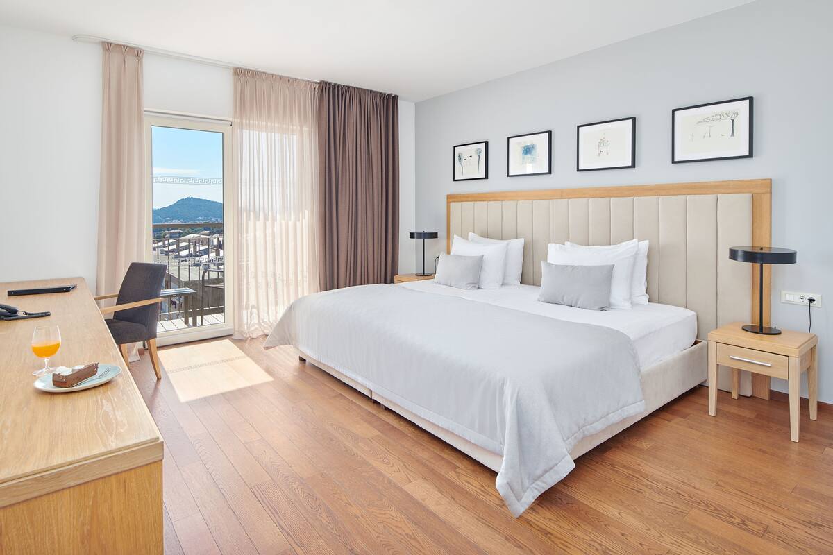 Habitación de hotel con una cama grande, un escritorio con comida y una bebida, una silla, una mesita de noche con una lámpara, y un balcón con vistas a la ciudad y a las montañas.