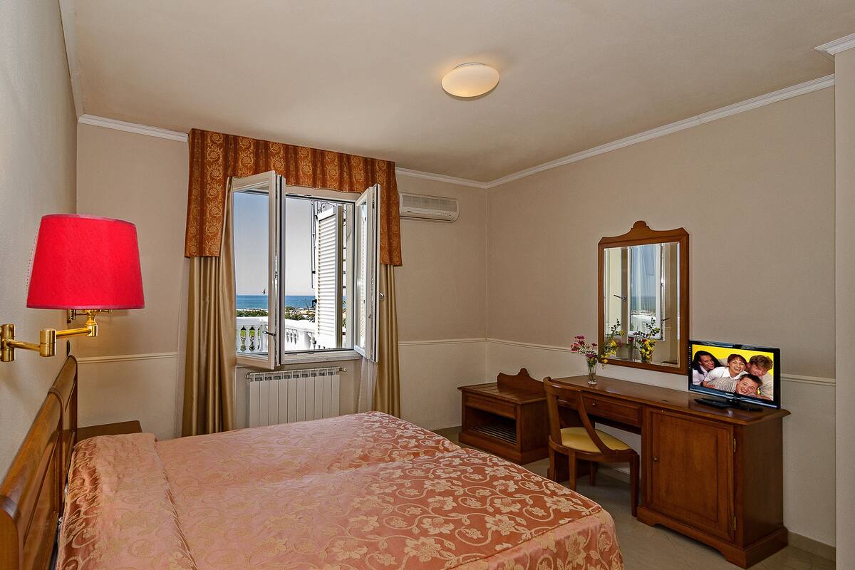 A hotel room with a bed, a desk, a chair, a television, a mirror, and a window with curtains.