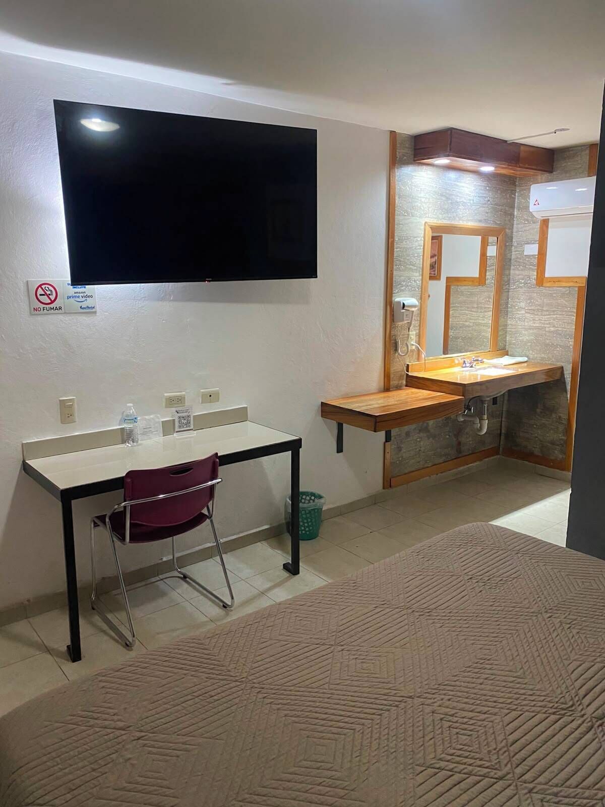 Una habitación con un televisor montado en la pared, un escritorio con una silla, un lavamanos con un espejo y una alfombra estampada.