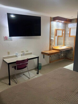 A room with a TV mounted on the wall, a desk with a chair, a sink with a mirror, and a patterned rug.