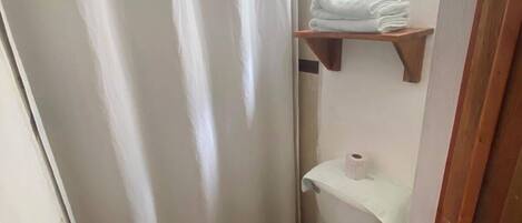 Un baño pequeño con cortina de ducha, inodoro, repisa con toallas y ventana sobre el inodoro.