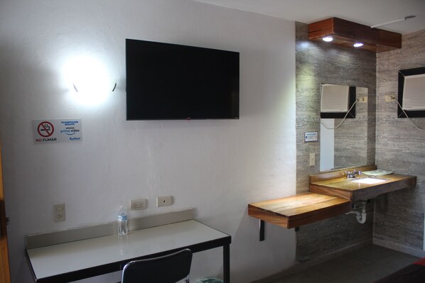 A modern bathroom with a flat-screen TV mounted on the wall, a wooden vanity with a sink, and a mirror.