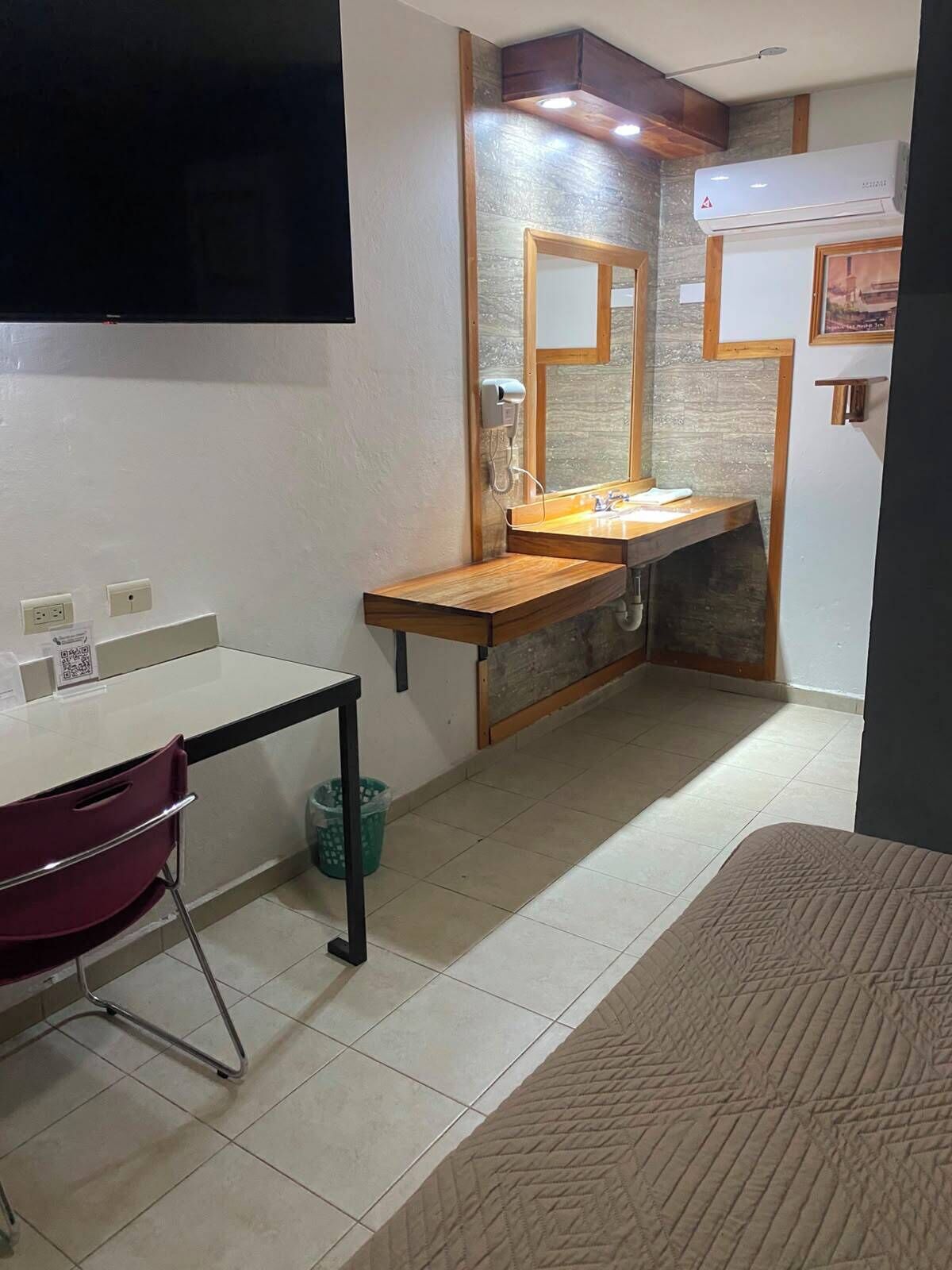 A compact hotel room with a wooden vanity, a mirror, a bed, and a television mounted on the wall.
