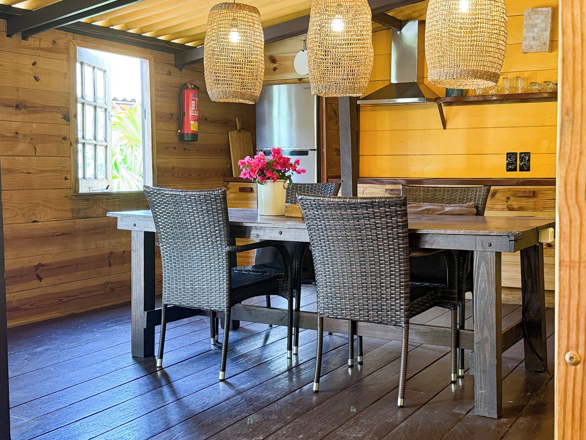 A wooden interior with a dining table, wicker chairs, and hanging wicker lamps.