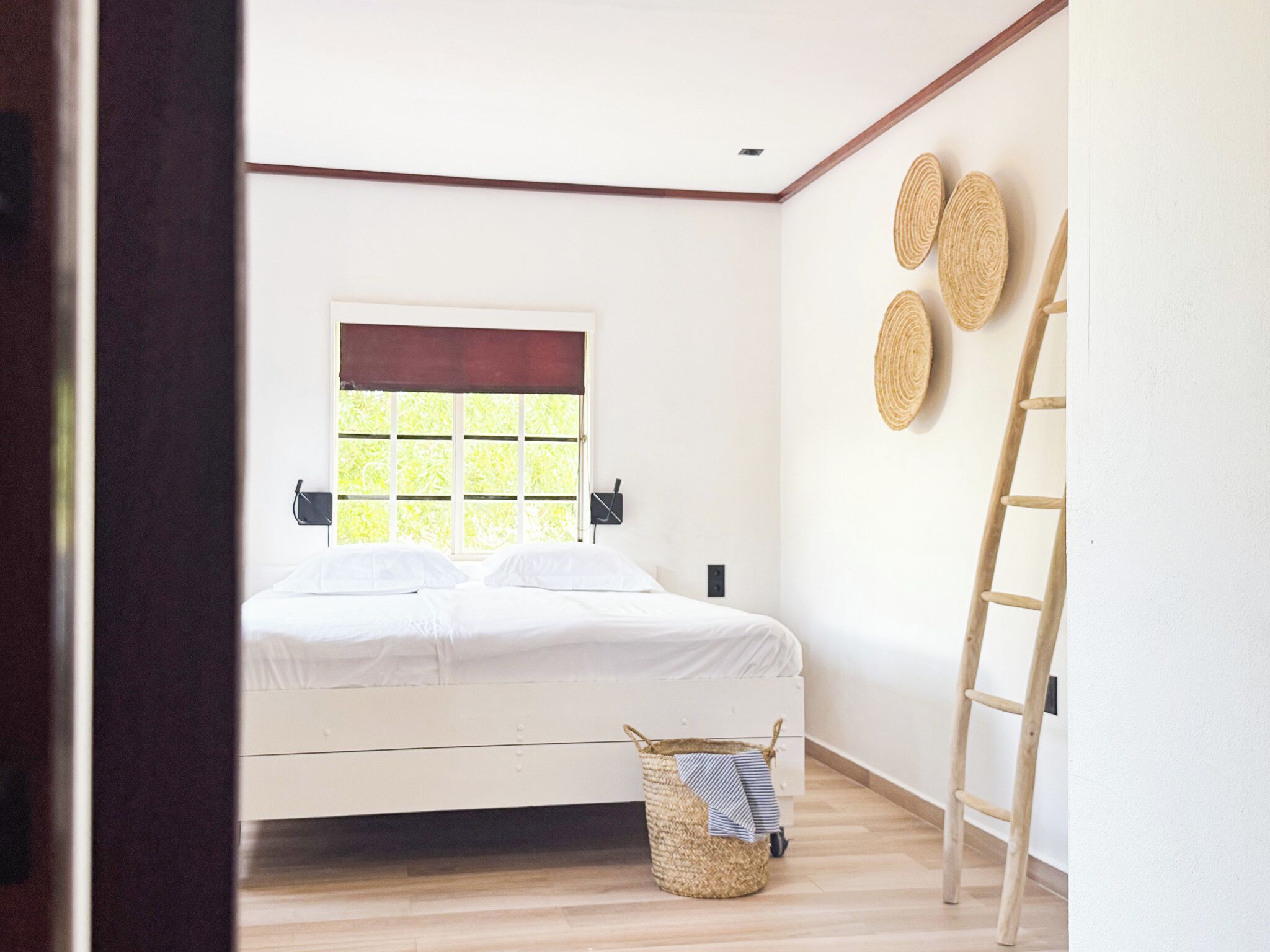 A bedroom with a bed, a window with blinds, a wall-mounted ladder, and a woven basket.