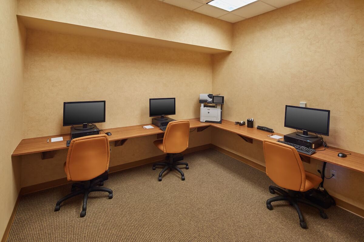 Habitación de hotel con un escritorio de madera, tres monitores de computadora y dos sillas de oficina.