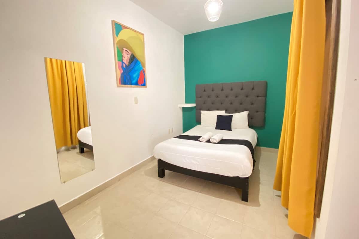 A hotel room with a bed, a painting on the wall, a mirror, and a light fixture.