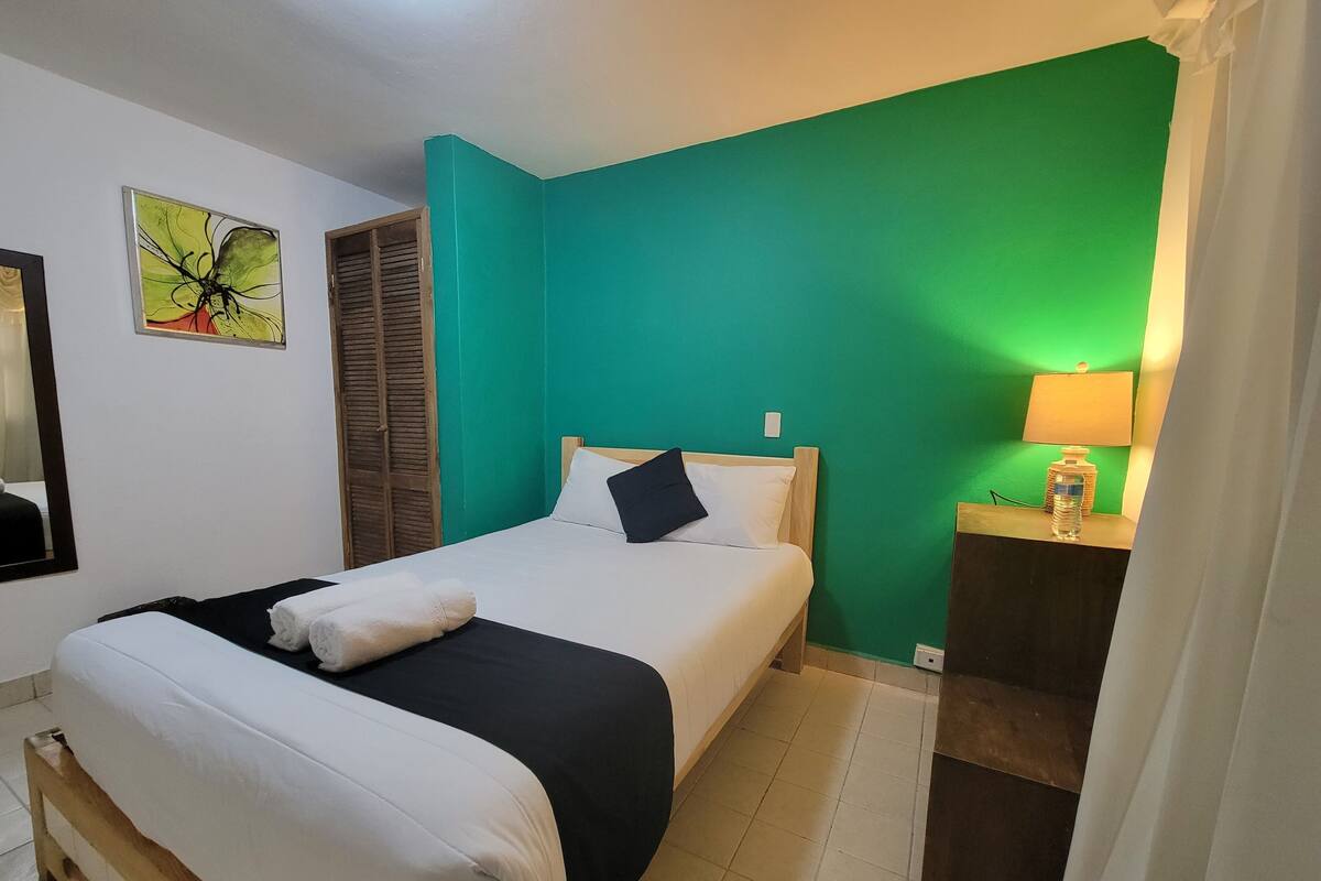 A hotel room with a bed, a nightstand with a lamp, a green accent wall, a wooden wardrobe, and a framed abstract painting.