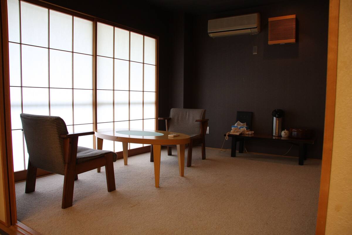 A traditional Japanese-style room with tatami mats, a low table, two chairs, and a sliding shoji screen.