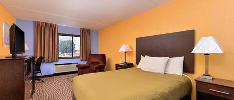 Habitación de hotel con una cama grande, un escritorio con computadora, una silla, una ventana con cortinas y una mesita de noche con lámpara.