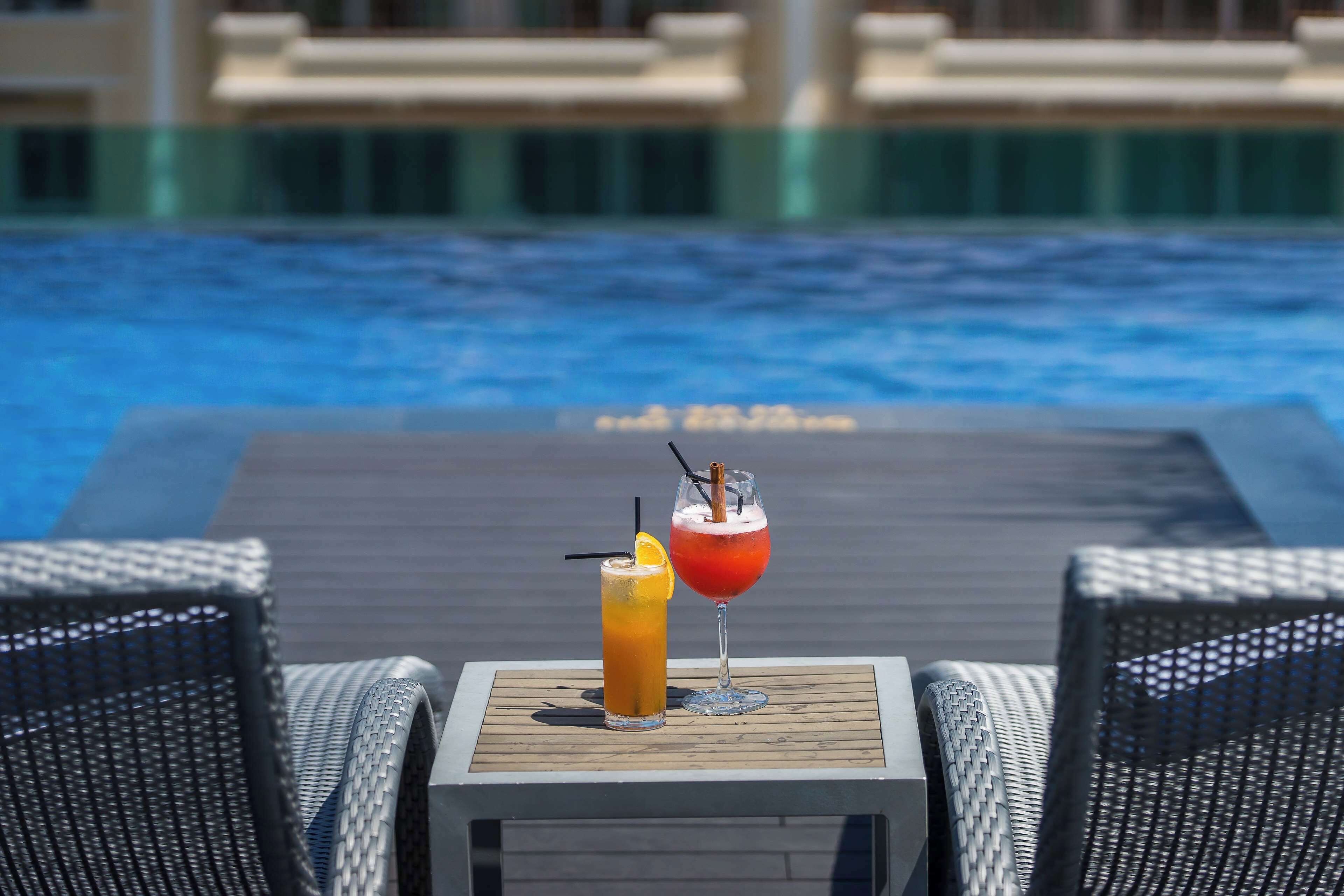 A poolside table with two drinks and wicker chairs.