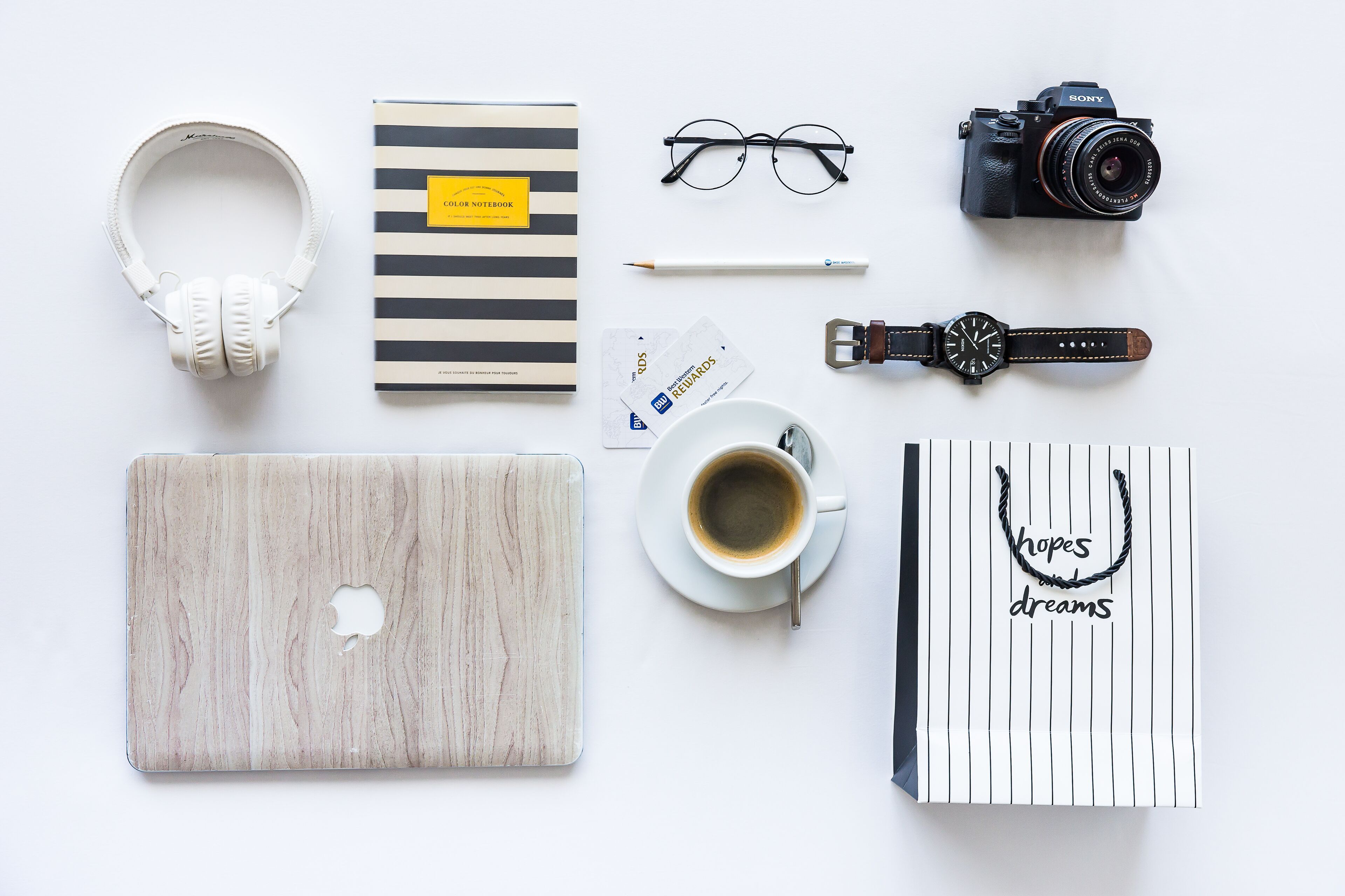Escritorio ordenado con una computadora portátil, audífonos, cámara, gafas, reloj, libreta, taza de café y una nota de motivación.